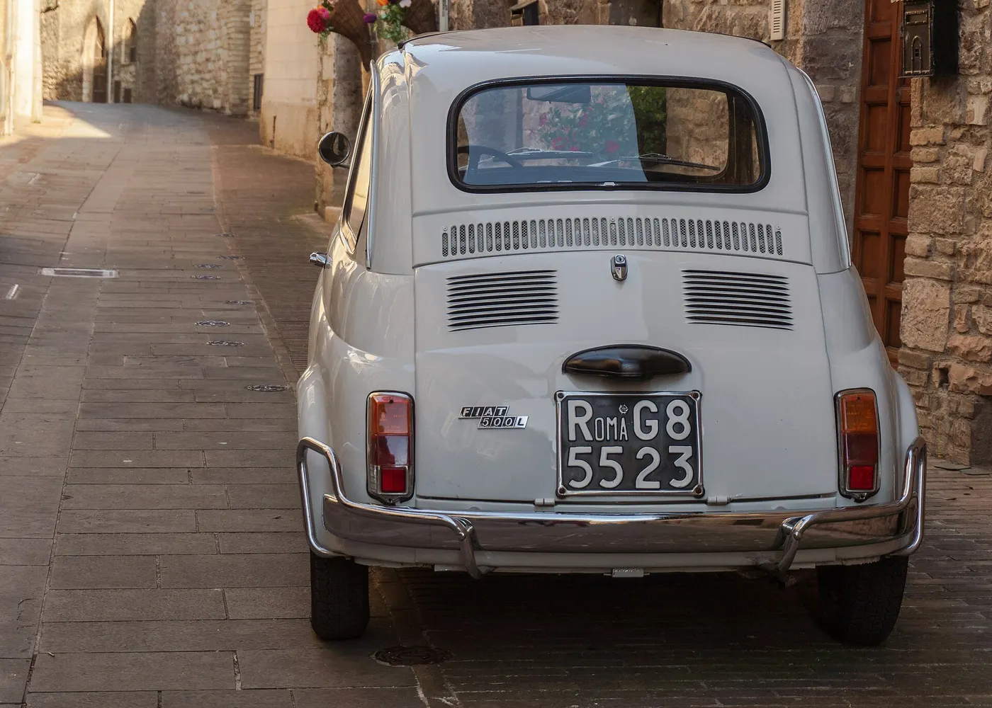 Una Fiat 500 L bianca degli anni Sessanta, vista da dietro, in una strada stretta